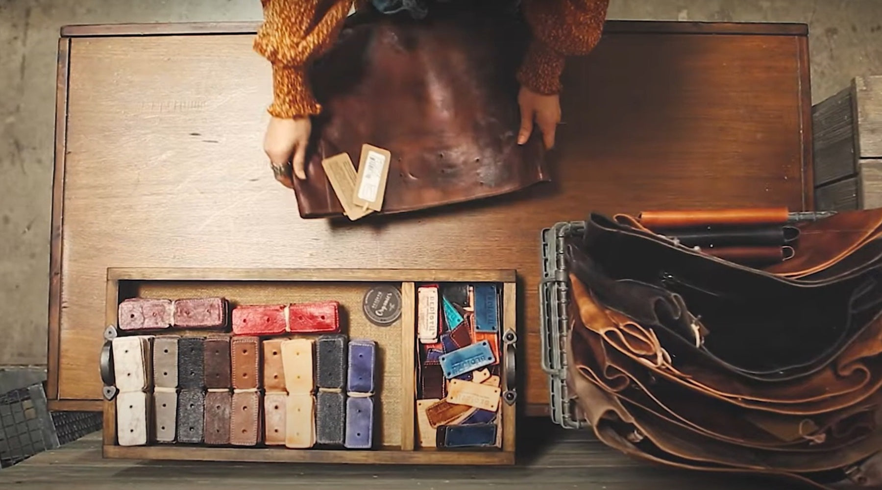 Person places a leather notebook next to a wooden box filled with colorful, labeled handmade soaps on a rustic table.