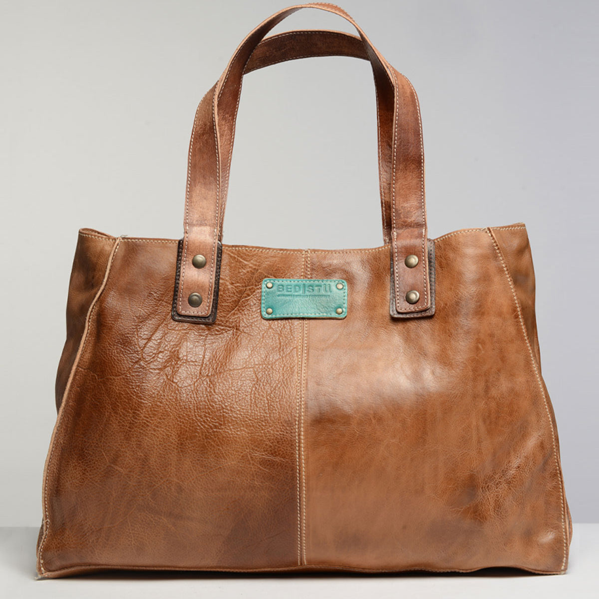 The Studio Arlington Body by Bed|Stü is a brown leather tote bag with dual handles and visible stitching, showcasing a teal metal brand plate on the front. This customizable bag lets you build your own by choosing details that match your personal style.
