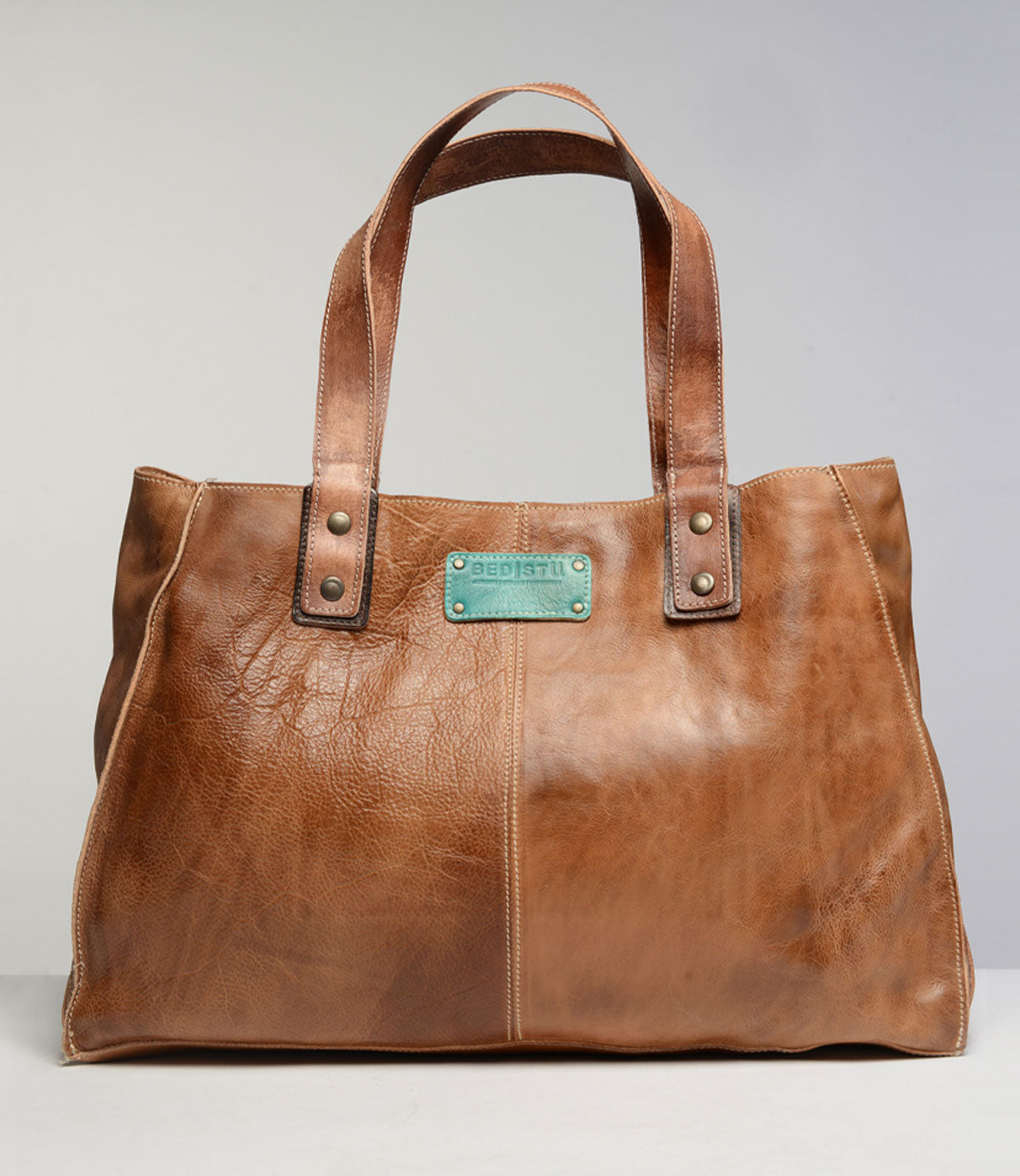 The Studio Arlington Body by Bed|Stü is a brown leather tote bag with dual handles and visible stitching, showcasing a teal metal brand plate on the front. This customizable bag lets you build your own by choosing details that match your personal style.