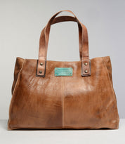 The Studio Arlington Body by Bed|Stü is a brown leather tote bag with dual handles and visible stitching, showcasing a teal metal brand plate on the front. This customizable bag lets you build your own by choosing details that match your personal style.