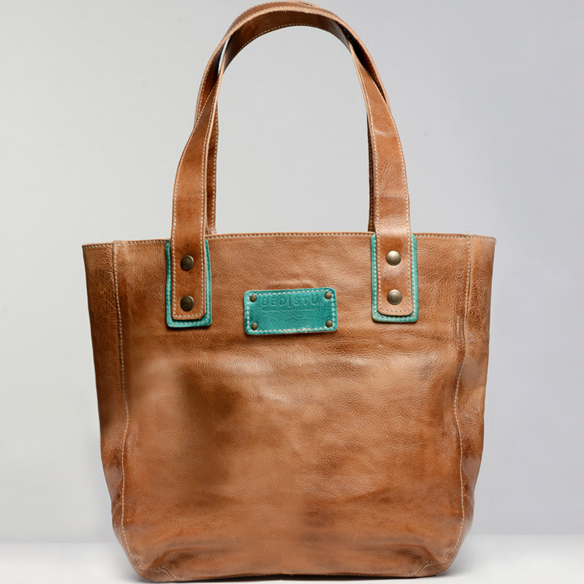 The Studio Gaviota Body by Bed|Stü is a brown leather tote bag featuring two handles and a teal-stitched rectangular patch near the top center, displayed against a plain gray background—a perfect personalized accessory to express your unique style.