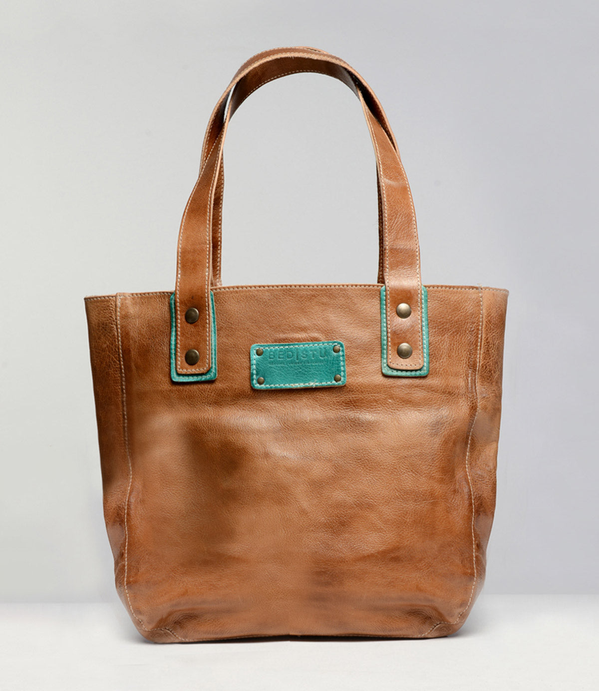 The Studio Gaviota Body by Bed|Stü is a brown leather tote bag featuring two handles and a teal-stitched rectangular patch near the top center, displayed against a plain gray background—a perfect personalized accessory to express your unique style.
