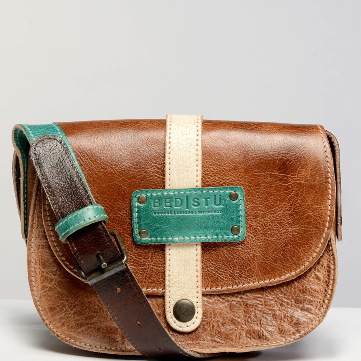 Introducing the Studio Shoreline Body crossbody bag by Bed|Stü: crafted in brown leather, featuring a beige vertical strap, a distinctive green accent patch with the "Bed|Stü" logo, and an adjustable shoulder strap edged in green. Set against a clean light backdrop, this customizable bag lets you express your personal style and create a look that’s all your own.