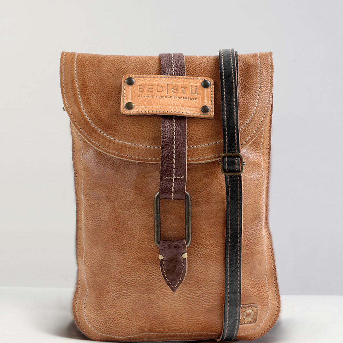 The Studio Sterling Body by Bed|Stü is a brown leather crossbody bag featuring a flap closure, striking buckle detail, and a contrasting black strap, showcased against a simple background—ideal for anyone seeking a customizable accessory to express their unique style.