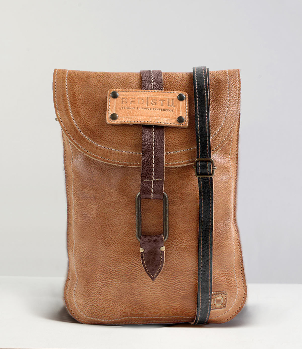 The Studio Sterling Body by Bed|Stü is a brown leather crossbody bag featuring a flap closure, striking buckle detail, and a contrasting black strap, showcased against a simple background—ideal for anyone seeking a customizable accessory to express their unique style.