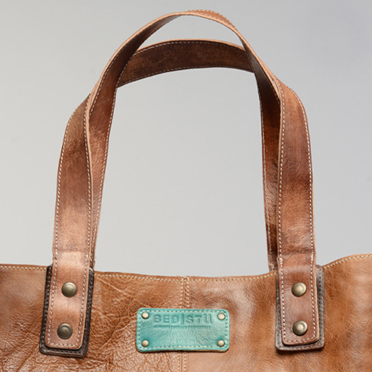 The Studio Short Shoulder Strap by Bed|Stü features brown leather, double handles, metal rivets, and a turquoise label with the "Bed|Stü" logo on the front—making it a perfect customizable bag for those seeking a unique personal touch.