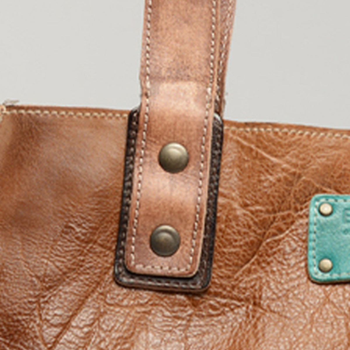 Close-up of the Bed|Stü Studio Strap Underlay, a brown leather customizable bag featuring a handle secured by two metal studs and a distinctive turquoise patch on the side.
