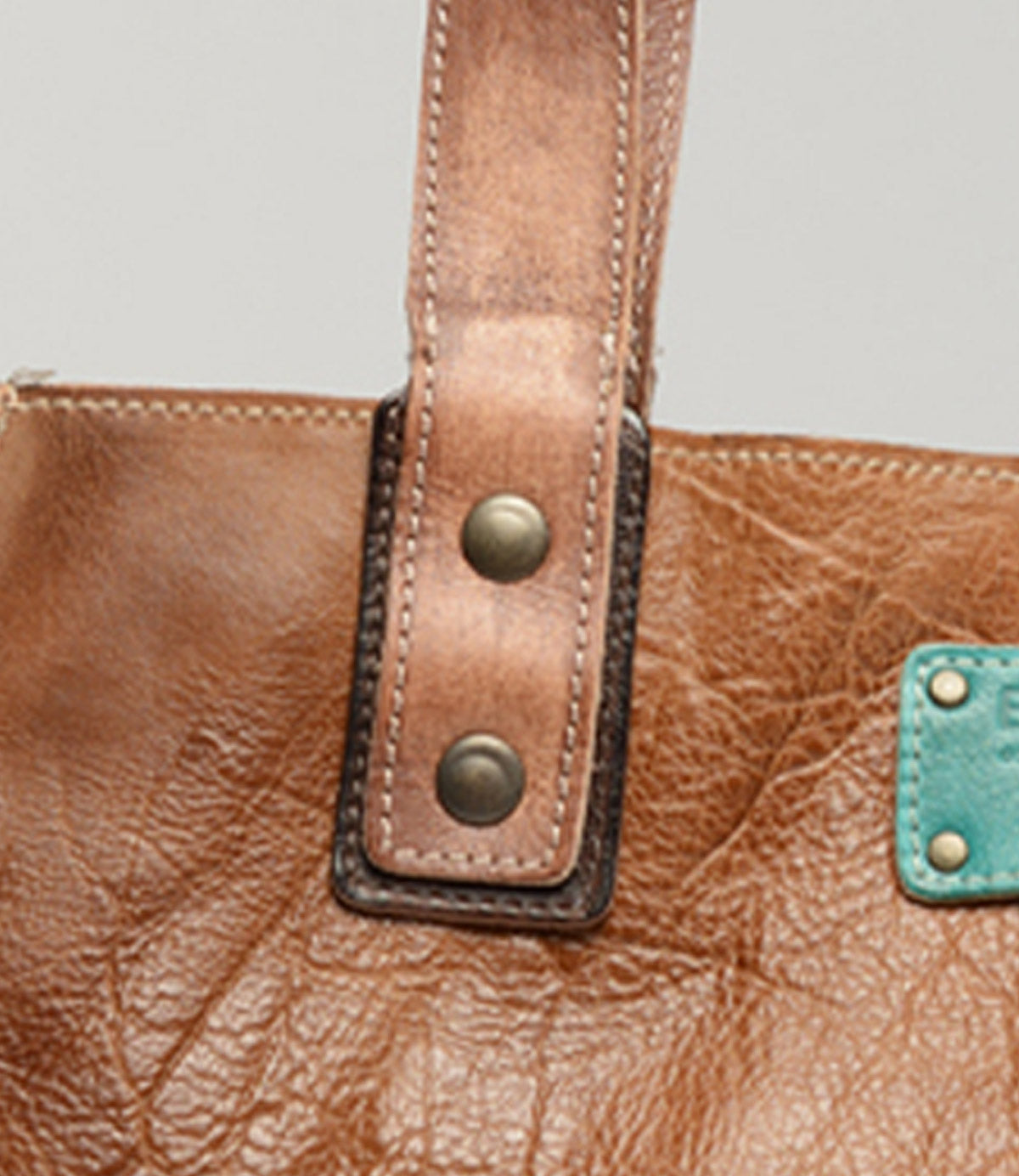 Close-up of the Bed|Stü Studio Strap Underlay, a brown leather customizable bag featuring a handle secured by two metal studs and a distinctive turquoise patch on the side.