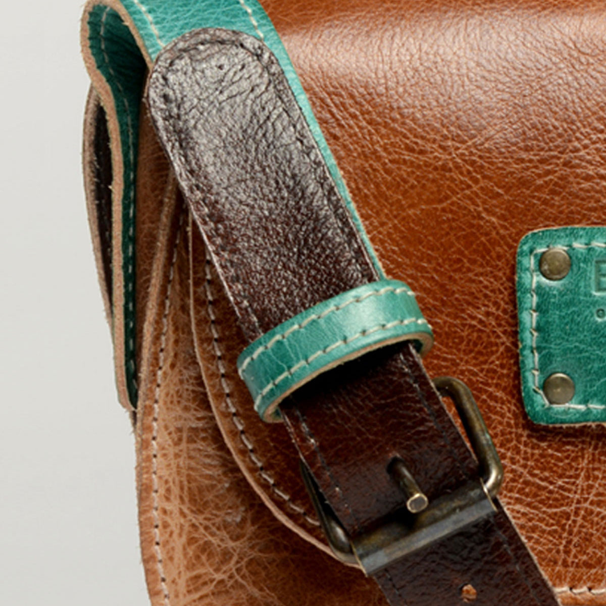 Close-up of the Bed|Stü Studio Buckle Strap—a brown leather bag with a green accent on the strap and a green patch on the flap, featuring a metal buckle. This stylish, customizable accessory lets you build your own bag for a truly personalized look.