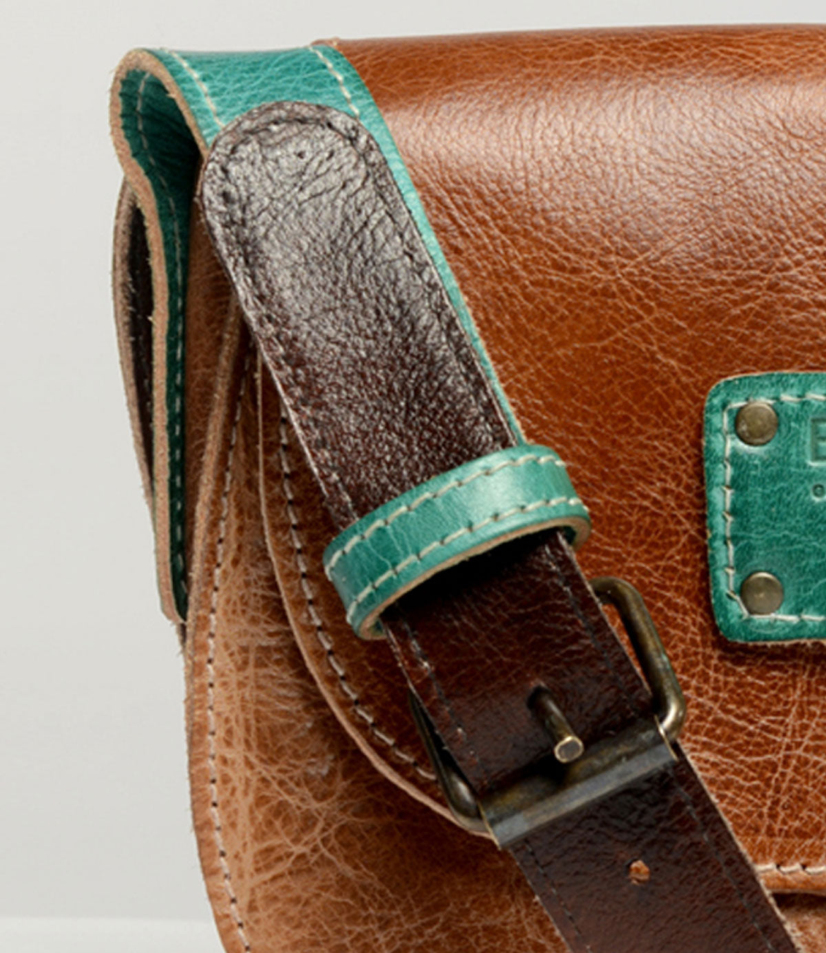 Close-up of the Bed|Stü Studio Buckle Strap—a brown leather bag with a green accent on the strap and a green patch on the flap, featuring a metal buckle. This stylish, customizable accessory lets you build your own bag for a truly personalized look.
