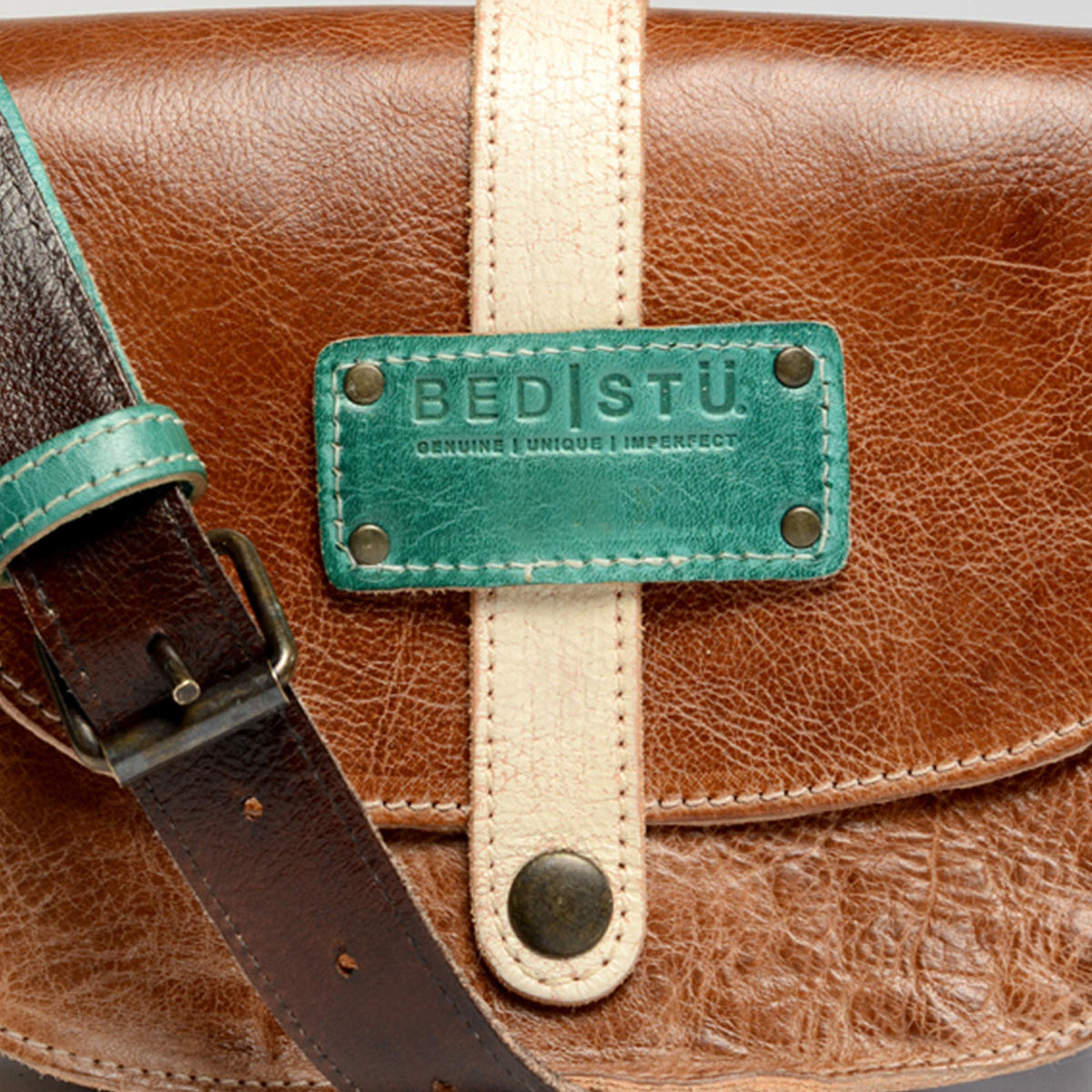 Close-up of a brown leather bag featuring Bed|Stü’s Studio Snap Strap—a white strap with a green label reading "BED|STÜ Genuine Unique Imperfect" and a dark brown buckle. This striking accessory is the perfect statement piece for those seeking to customize their bag and express individuality with their own unique design.