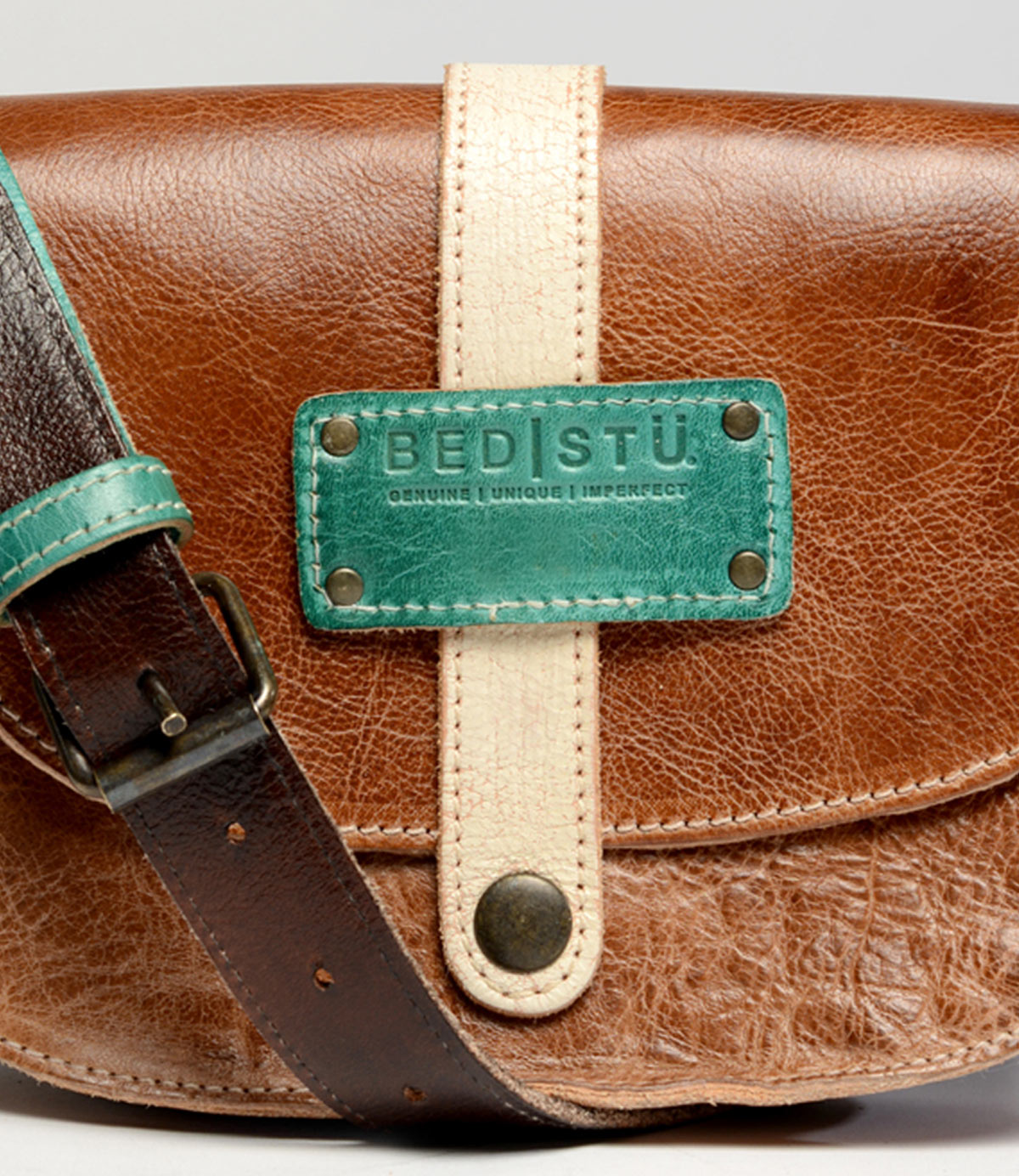 Close-up of a brown leather bag featuring Bed|Stü’s Studio Snap Strap—a white strap with a green label reading "BED|STÜ Genuine Unique Imperfect" and a dark brown buckle. This striking accessory is the perfect statement piece for those seeking to customize their bag and express individuality with their own unique design.