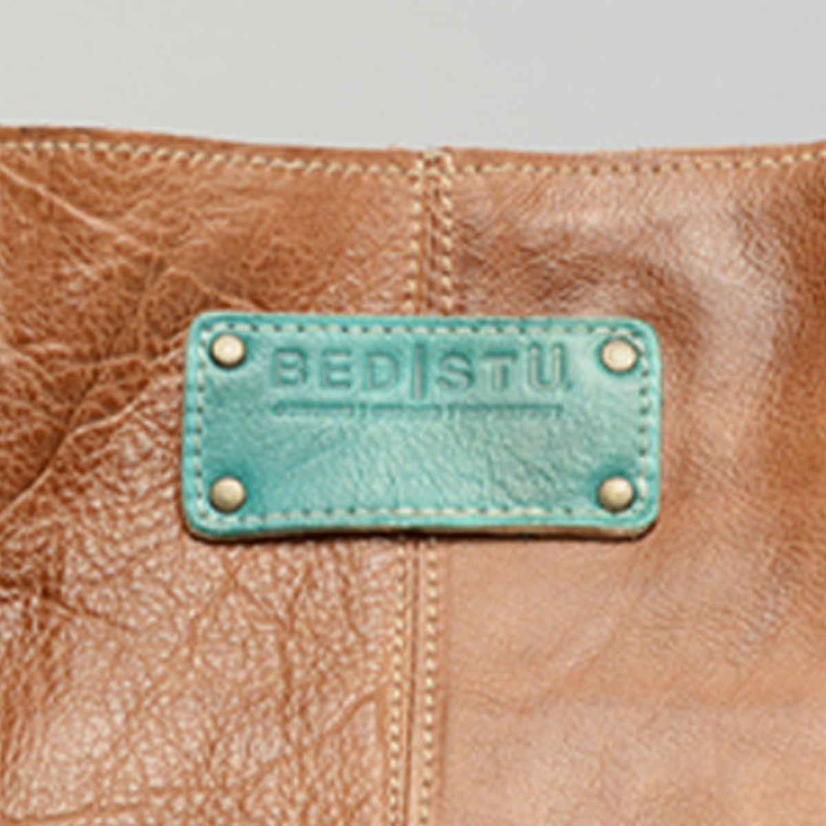 Close-up of a brown leather Studio Logo Patch bag by Bed|Stü, featuring a teal patch with the embossed brand name secured by four metal rivets.