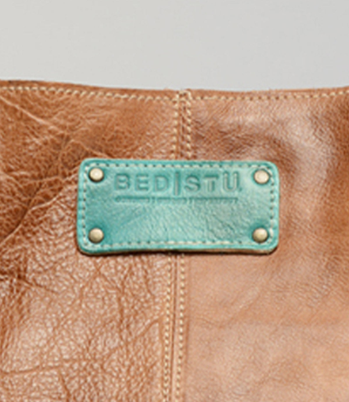 Close-up of a brown leather Studio Logo Patch bag by Bed|Stü, featuring a teal patch with the embossed brand name secured by four metal rivets.