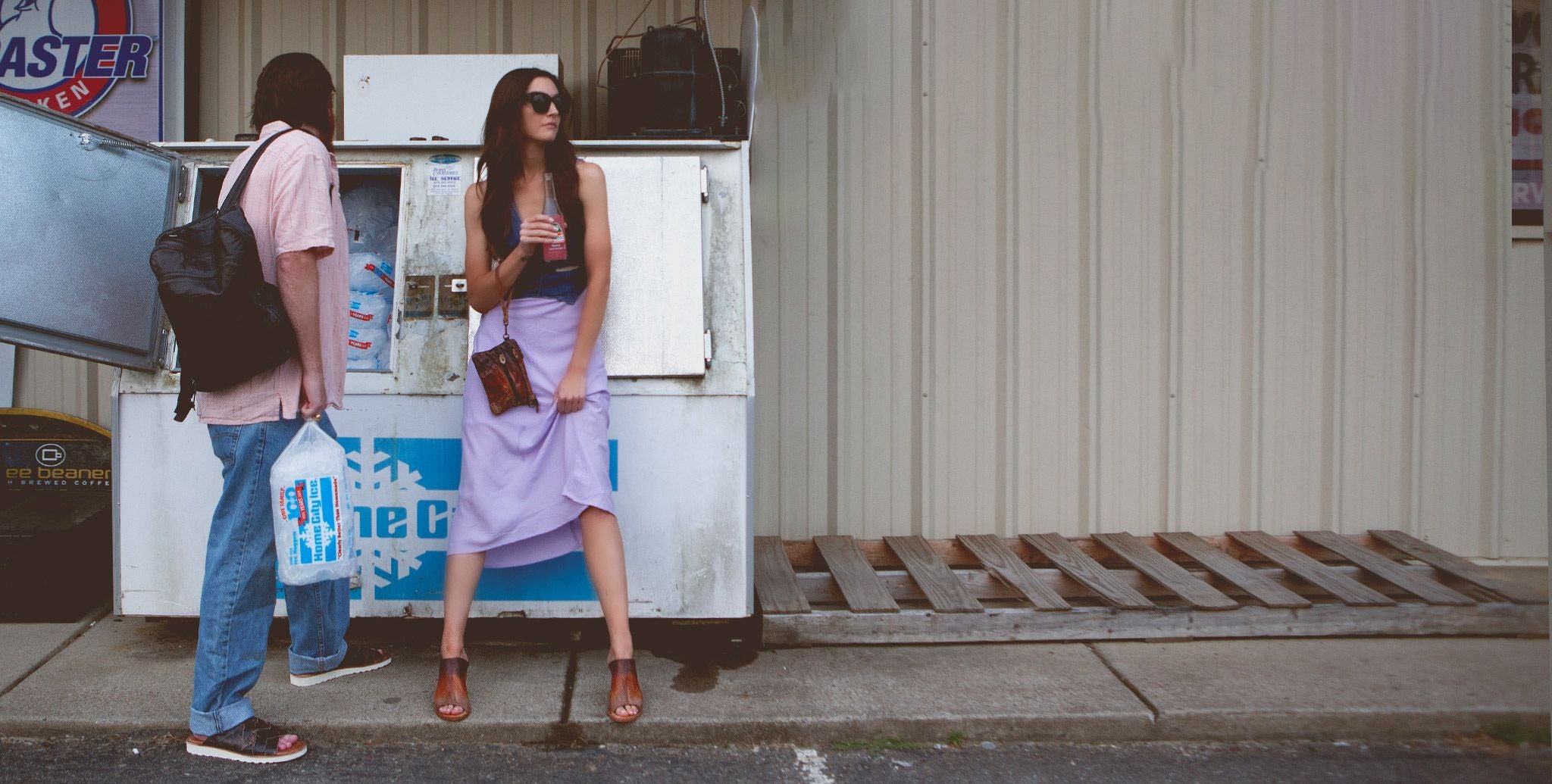 A women and man are standing in a convenience store parking lots by an ice machine.  He is in black leather snadls, denim pants and a coral button up shirt carrying a black backpack on his shoulder and holding a bag of ice. She is wearing a purple skirt and denim vest drink a pink soda from a glass bottle leaning against the ice machine. 