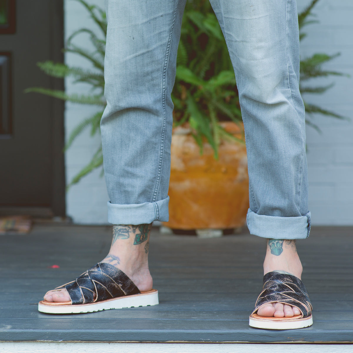 Wearing rolled-up light jeans, a person stands on a porch in the Bed|Stü Abraham Light sandals, which feature a woven leather upper and lightweight outsole for all-day comfort; tattoos are visible on their feet and ankles, with a potted fern in the background.