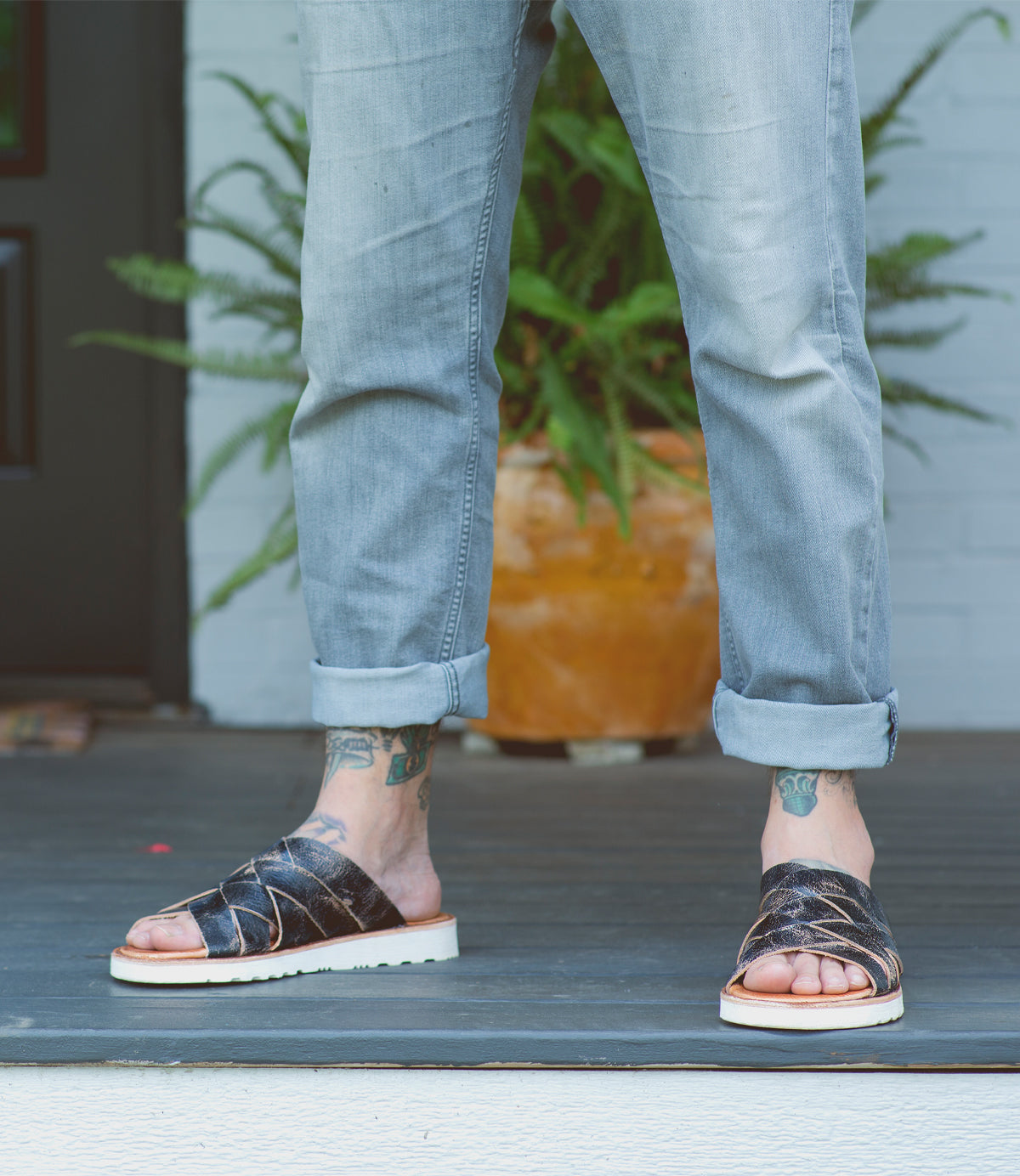 Wearing rolled-up light jeans, a person stands on a porch in the Bed|Stü Abraham Light sandals, which feature a woven leather upper and lightweight outsole for all-day comfort; tattoos are visible on their feet and ankles, with a potted fern in the background.
