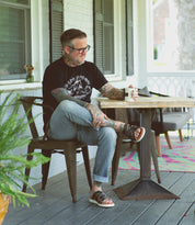A man with tattoos and glasses sits on a porch chair, holding a coffee mug with one leg crossed over the other. He wears a black t-shirt, rolled-up jeans, and Bed|Stü's Abraham Light sandals—crafted with a woven leather upper for casual style and all-day comfort.