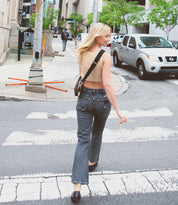 A woman with blonde hair crosses a city street at a crosswalk, holding a small object and wearing the stylish Bed|Stü Abundance crossbody bag, crafted from sustainable reclaimed leather, with cars and buildings visible in the background.