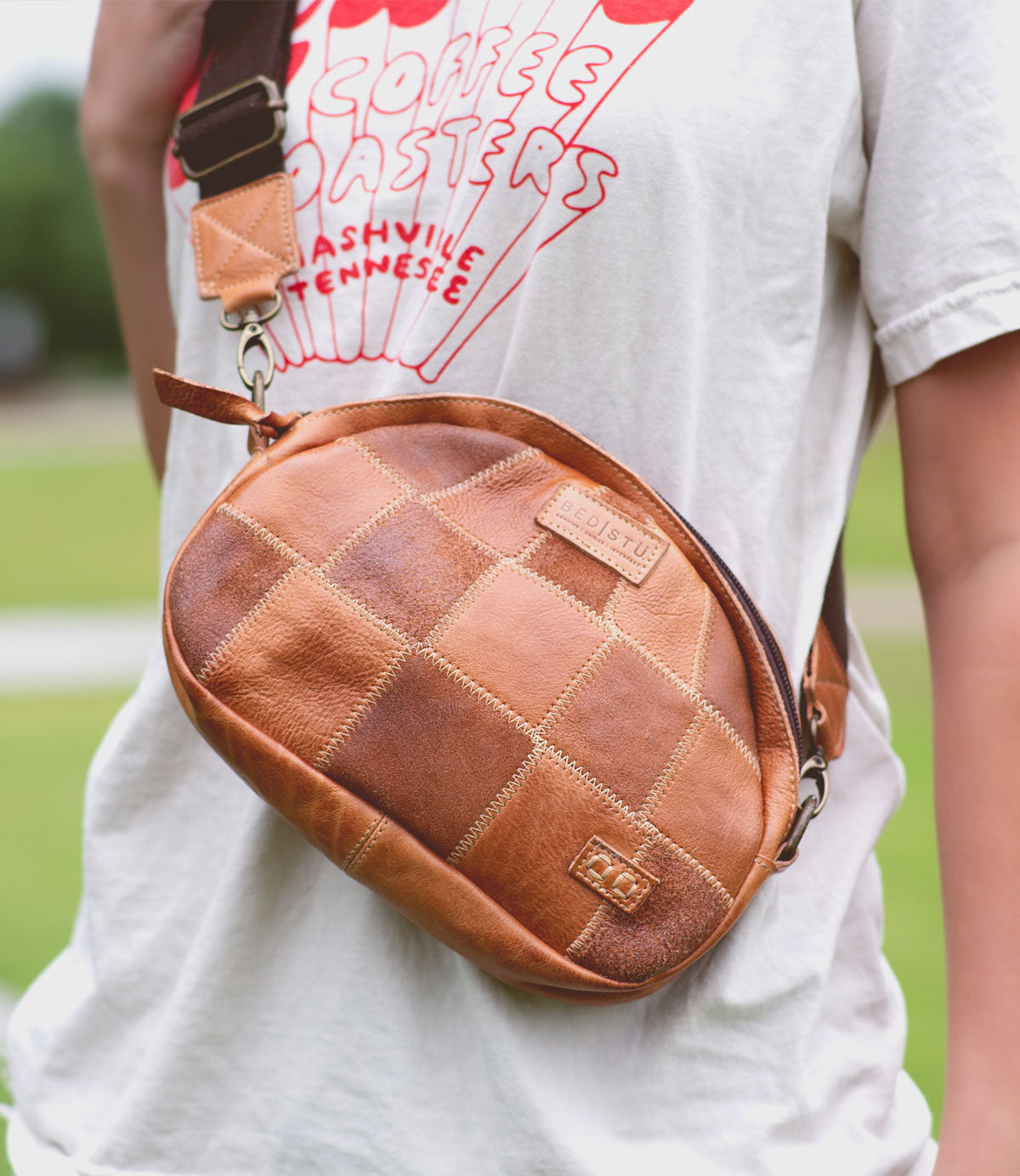 A person wearing the Bed|Stü Abundance patchwork brown crossbody bag, crafted from reclaimed leather, over a white graphic t-shirt with red text in an outdoor setting, showcasing a sustainable style.