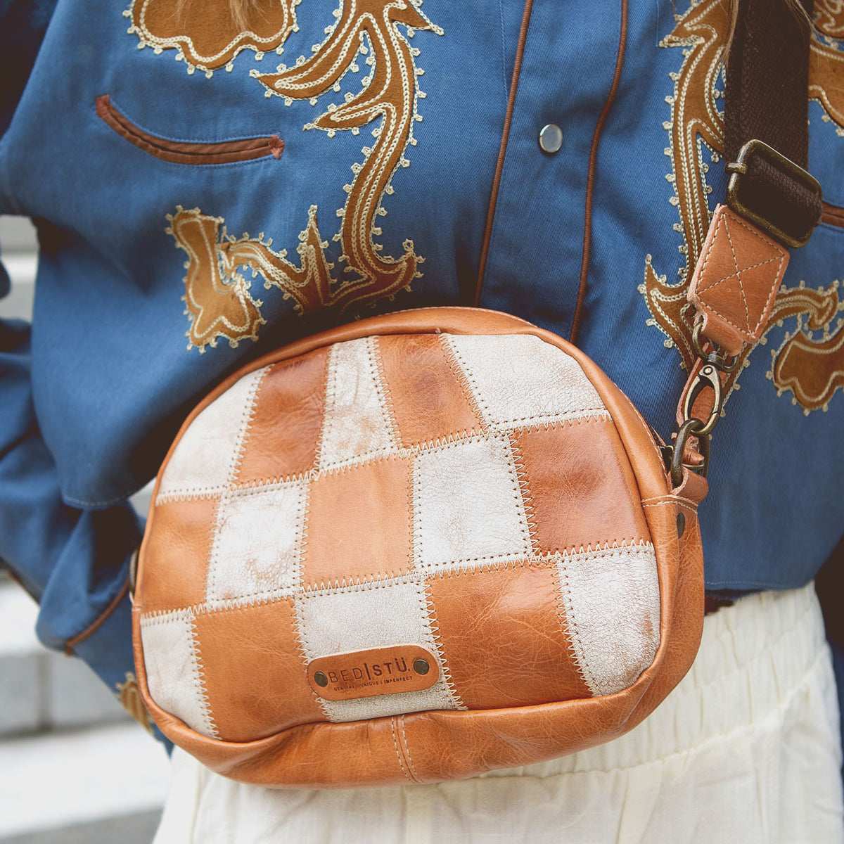 A person wearing a blue embroidered shirt, white skirt, and carrying the Bed|Stü Abundance crossbody bag—a small brown-and-white checkered bag crafted from reclaimed leather with a visible brand label.