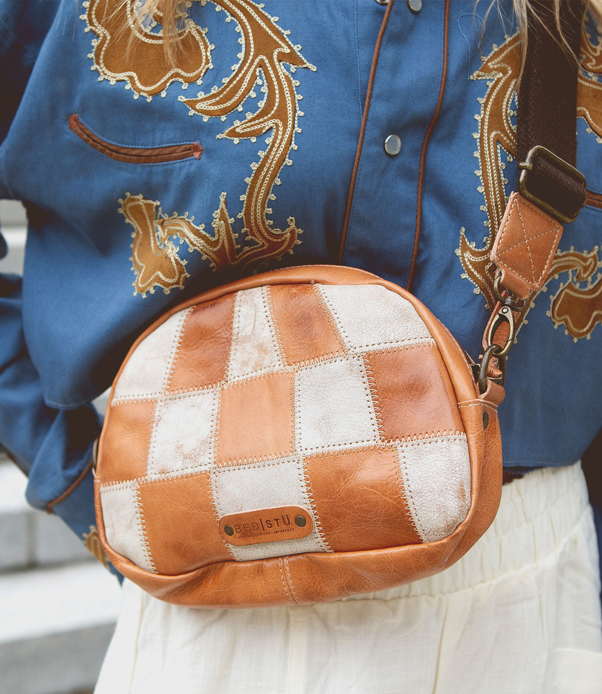 A person wearing a blue embroidered shirt, white skirt, and carrying the Bed|Stü Abundance crossbody bag—a small brown-and-white checkered bag crafted from reclaimed leather with a visible brand label.