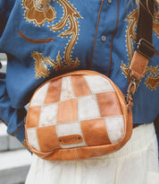 A person wearing a blue embroidered shirt, white skirt, and carrying the Bed|Stü Abundance crossbody bag—a small brown-and-white checkered bag crafted from reclaimed leather with a visible brand label.