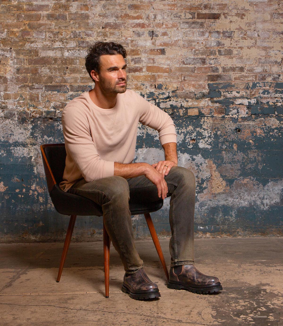A man with dark hair sits on a chair against a worn brick wall, wearing a light long-sleeve shirt and gray jeans, and finishing his look with the Bed|Stü Accelerate brown Chelsea boots featuring a lug outsole; he gazes to the side with a neutral expression that brings a modern edge to the scene.