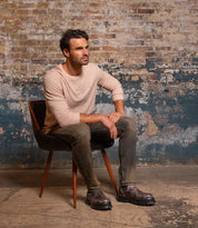 A man with dark hair sits on a chair against a worn brick wall, wearing a light long-sleeve shirt and gray jeans, and finishing his look with the Bed|Stü Accelerate brown Chelsea boots featuring a lug outsole; he gazes to the side with a neutral expression that brings a modern edge to the scene.