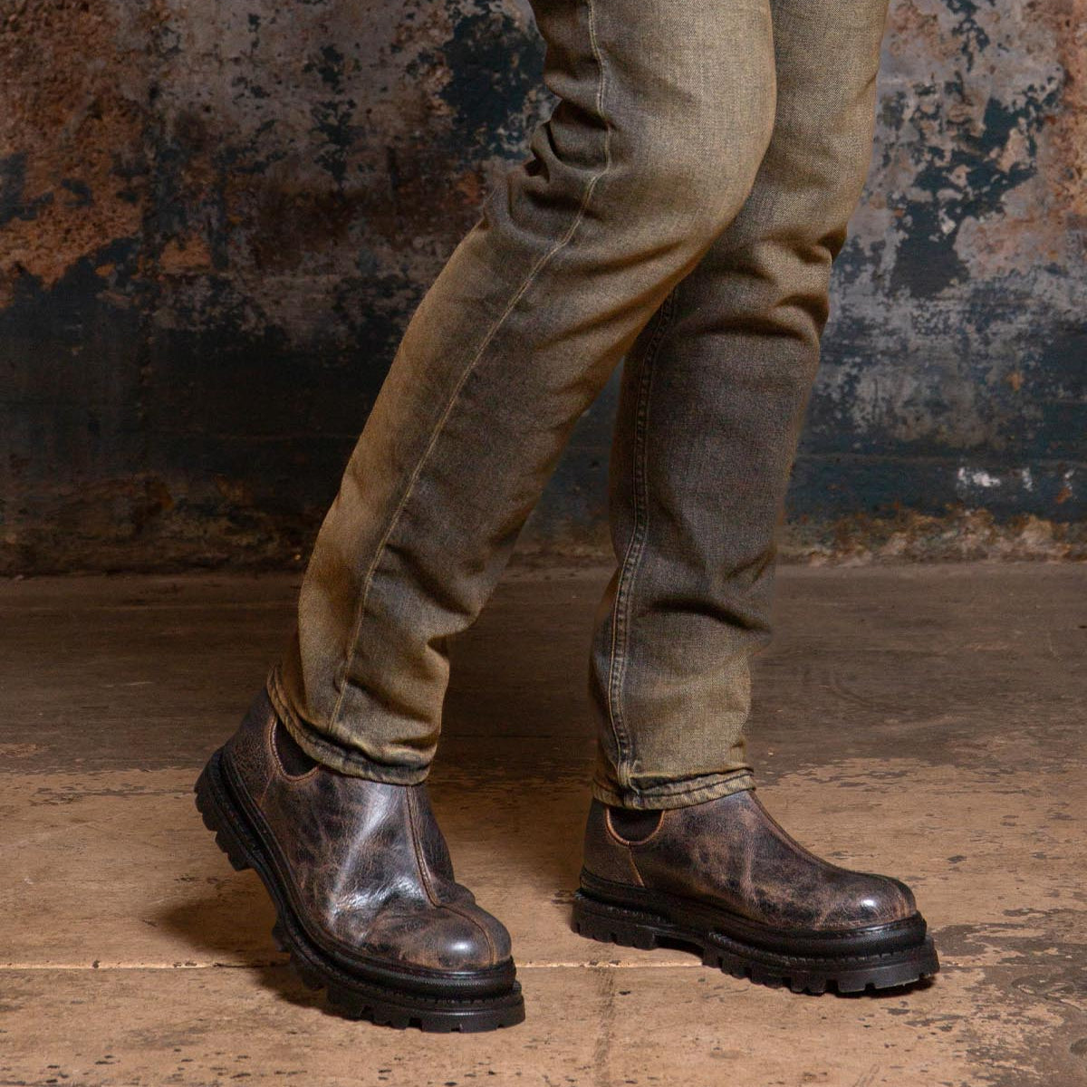 Wearing the Accelerate distressed jeans and Bed|Stü’s dark Chelsea boots with a lug outsole, a person stands on a concrete floor against a textured, worn wall, giving the look a modern edge.