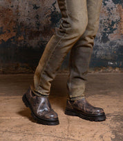Wearing the Accelerate distressed jeans and Bed|Stü’s dark Chelsea boots with a lug outsole, a person stands on a concrete floor against a textured, worn wall, giving the look a modern edge.
