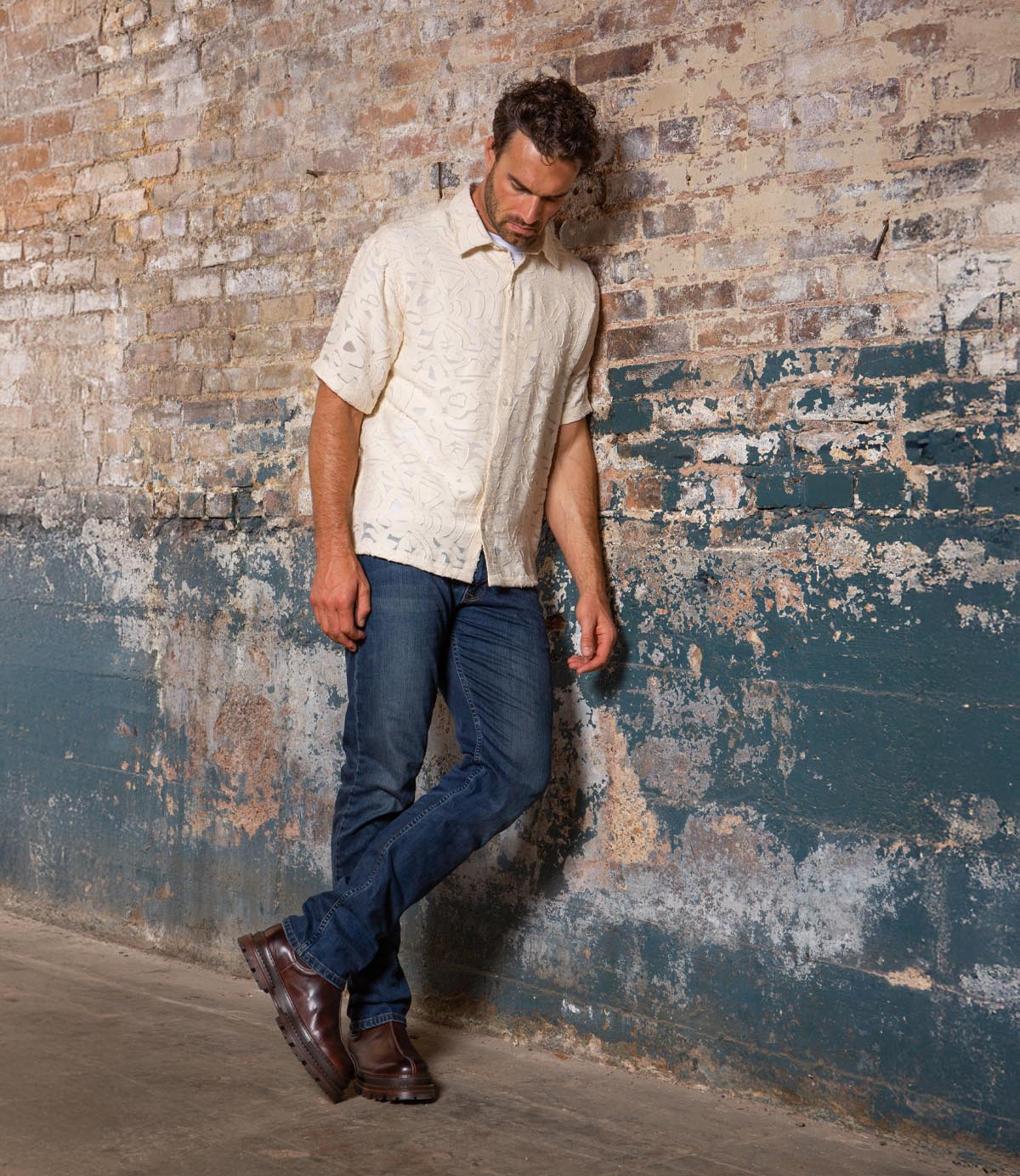 Wearing the Bed|Stü Accelerate brown Chelsea boots with a lug outsole, this man in a cream short-sleeve shirt and blue jeans stands against a worn brick wall, looking down with one foot crossed over the other—giving his classic style a modern edge.