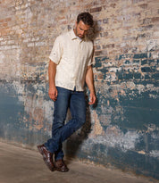 Wearing the Bed|Stü Accelerate brown Chelsea boots with a lug outsole, this man in a cream short-sleeve shirt and blue jeans stands against a worn brick wall, looking down with one foot crossed over the other—giving his classic style a modern edge.