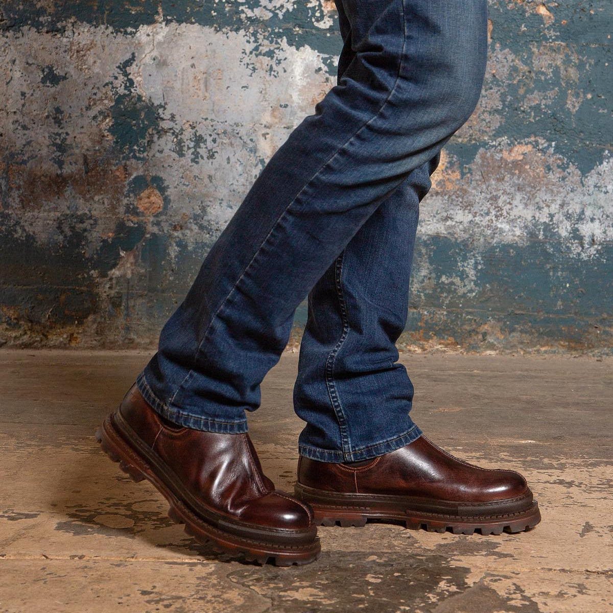 A person wearing the Bed|Stü Accelerate dark brown Chelsea boots with a lug outsole and blue jeans stands on a worn concrete floor, set against a distressed blue and brown wall that adds a modern edge to the scene.