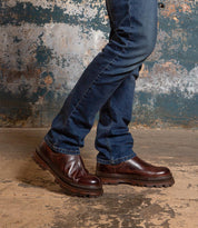 A person wearing the Bed|Stü Accelerate dark brown Chelsea boots with a lug outsole and blue jeans stands on a worn concrete floor, set against a distressed blue and brown wall that adds a modern edge to the scene.