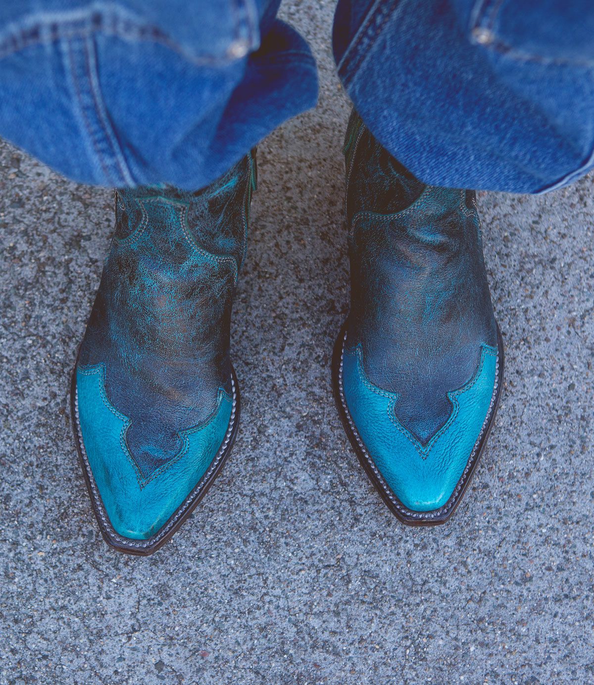 A person wearing the Bed|Stü Ace pointed turquoise and black Western boots, crafted from full-grain leather, stands on a rough gray concrete surface with blue jeans partially covering the boots—a nod to traditional boot making.
