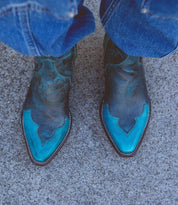 A person wearing the Bed|Stü Ace pointed turquoise and black Western boots, crafted from full-grain leather, stands on a rough gray concrete surface with blue jeans partially covering the boots—a nod to traditional boot making.