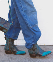 A person walks on the pavement wearing Bed|Stü's Ace teal-and-black Western boots, expertly crafted from full-grain leather using traditional boot making techniques, paired with a matching teal-and-black bag.