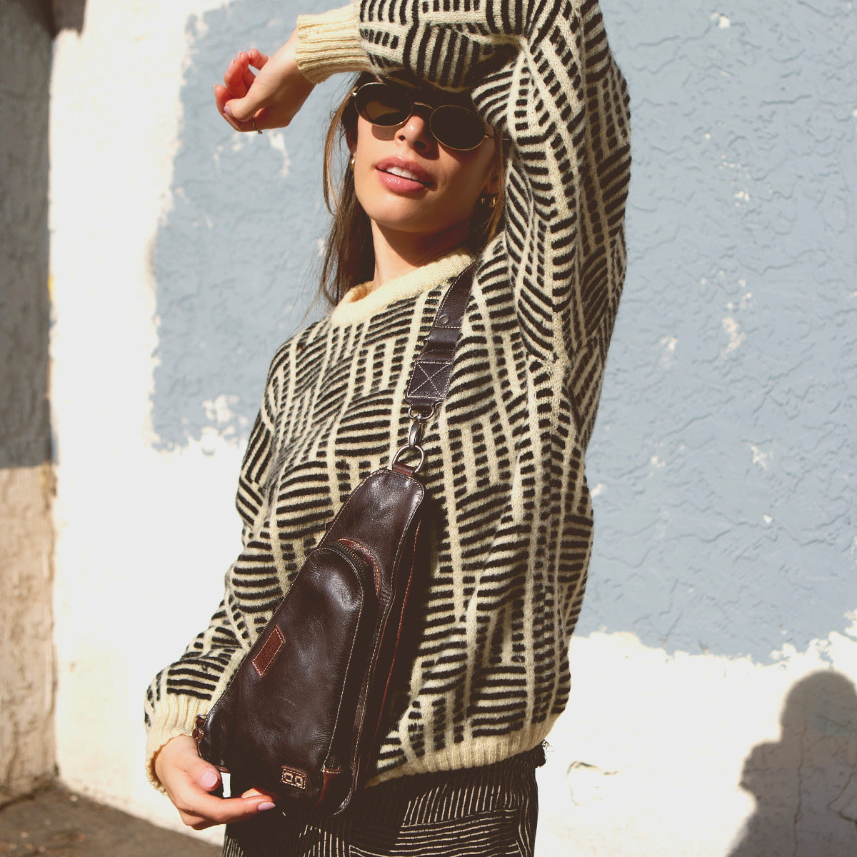 A woman in sunglasses stands outside, dressed in a patterned sweater and striped pants, wearing the Bed|Stü Adept—a compact sling crossbody bag crafted from vegetable-tanned leather with an adjustable strap—one arm raised to shield her face from the sun.