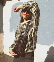 A woman in sunglasses stands outside, dressed in a patterned sweater and striped pants, wearing the Bed|Stü Adept—a compact sling crossbody bag crafted from vegetable-tanned leather with an adjustable strap—one arm raised to shield her face from the sun.