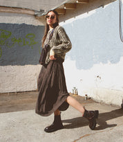 A woman wearing sunglasses, a patterned sweater, a pleated skirt, and black boots walks outside along a sunlit concrete area beside a painted wall with graffiti, carrying the Bed|Stü Adept—a compact sling with an adjustable strap crafted from vegetable-tanned leather.