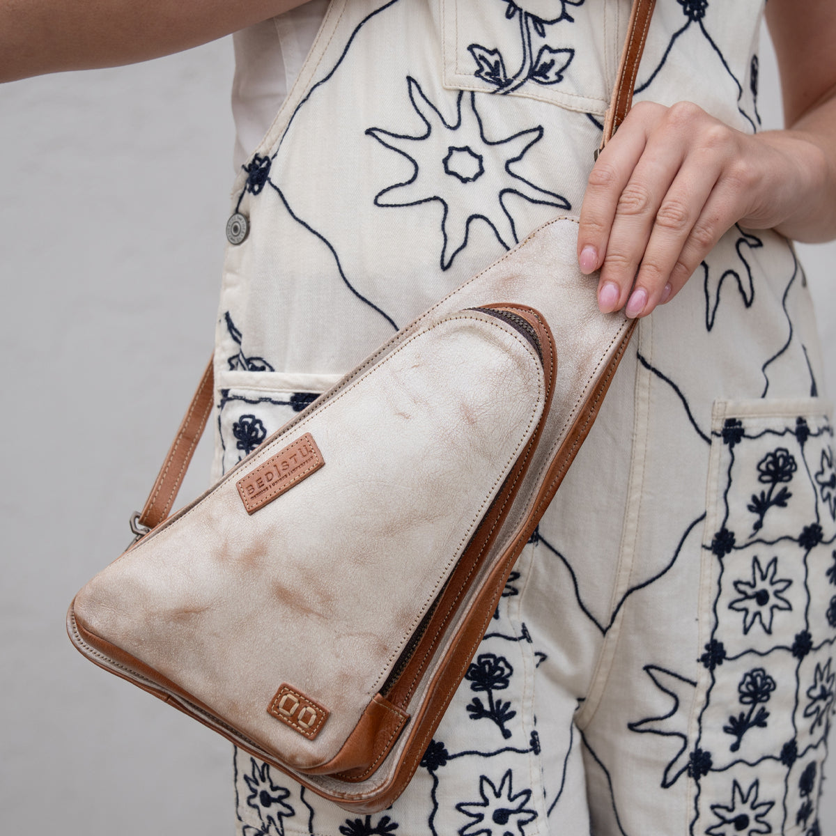 A person wearing a cream jumpsuit with dark floral embroidery holds the Bed|Stü Adept, a tan and cream triangular compact sling crossbody bag crafted from vegetable-tanned leather, featuring an adjustable strap and a visible brand patch.