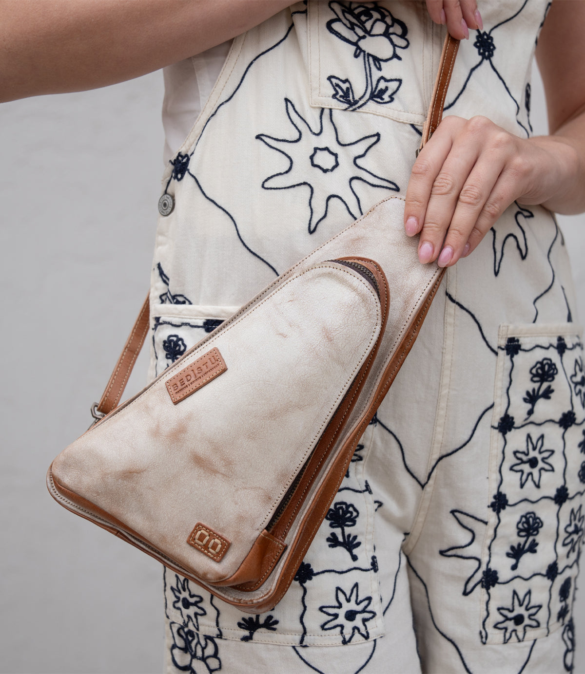A person wearing a cream jumpsuit with dark floral embroidery holds the Bed|Stü Adept, a tan and cream triangular compact sling crossbody bag crafted from vegetable-tanned leather, featuring an adjustable strap and a visible brand patch.