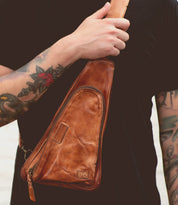 A person with a tattooed arm wears the Bed|Stü Adept sling bag, crafted from brown vegetable-tanned leather, across their chest. The compact bag features an adjustable strap and is paired with a black shirt.