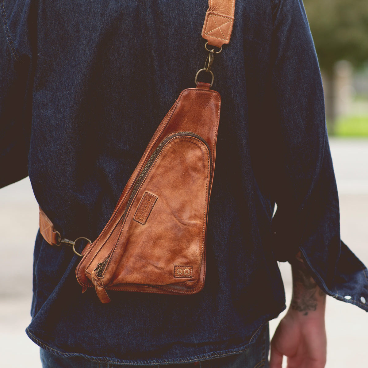 A person wearing the Adept crossbody sling bag by Bed|Stü, crafted from brown vegetable-tanned leather and featuring an adjustable strap, over a dark denim shirt, photographed from behind.