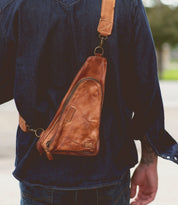 A person wearing the Adept crossbody sling bag by Bed|Stü, crafted from brown vegetable-tanned leather and featuring an adjustable strap, over a dark denim shirt, photographed from behind.