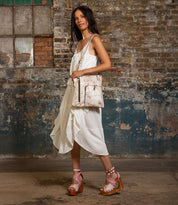 A woman in a white dress stands indoors against a distressed brick wall, wearing brown platform sandals and carrying the Bed|Stü Aiken full-grain leather bag with an adjustable strap.