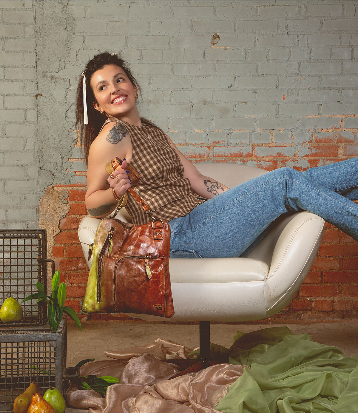 A woman sits sideways on a white chair, smiling and holding the Bed|Stü Aiken full-grain leather bag with an adjustable strap, which contains a pear. Pears and green fabric are arranged on the floor next to black metal crates against a brick wall.