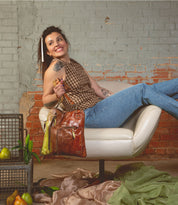 A woman sits sideways on a white chair, smiling and holding the Bed|Stü Aiken full-grain leather bag with an adjustable strap, which contains a pear. Pears and green fabric are arranged on the floor next to black metal crates against a brick wall.