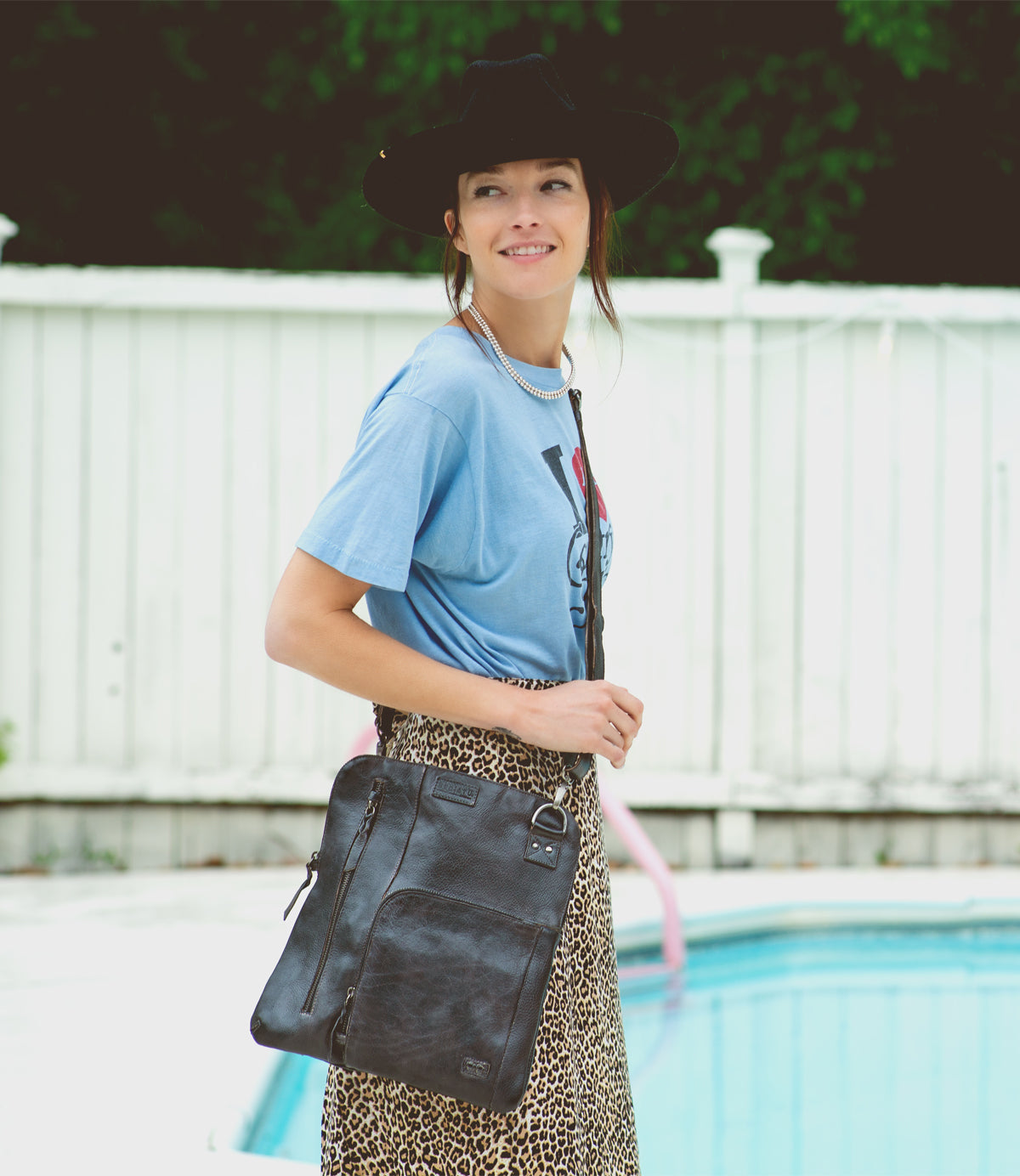 A woman stands by a pool, smiling and carrying the Bed|Stü Aiken full-grain leather bag with an adjustable strap, paired with a blue T-shirt, leopard print skirt, and black hat.